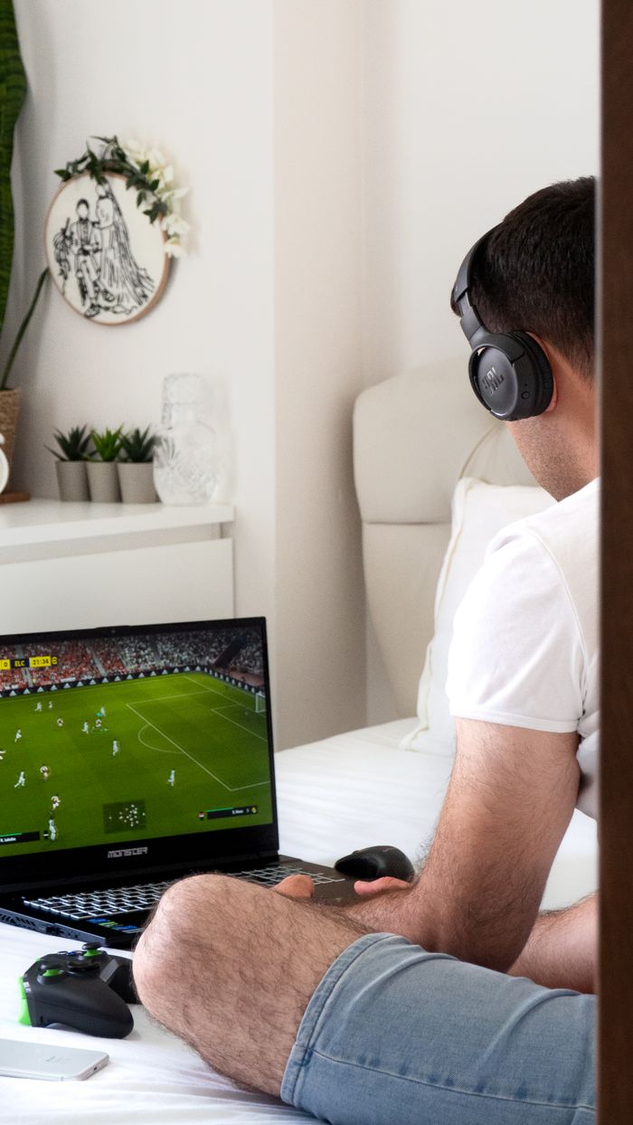Man sitting on bed gaming with headphones and laptop in a bright room.