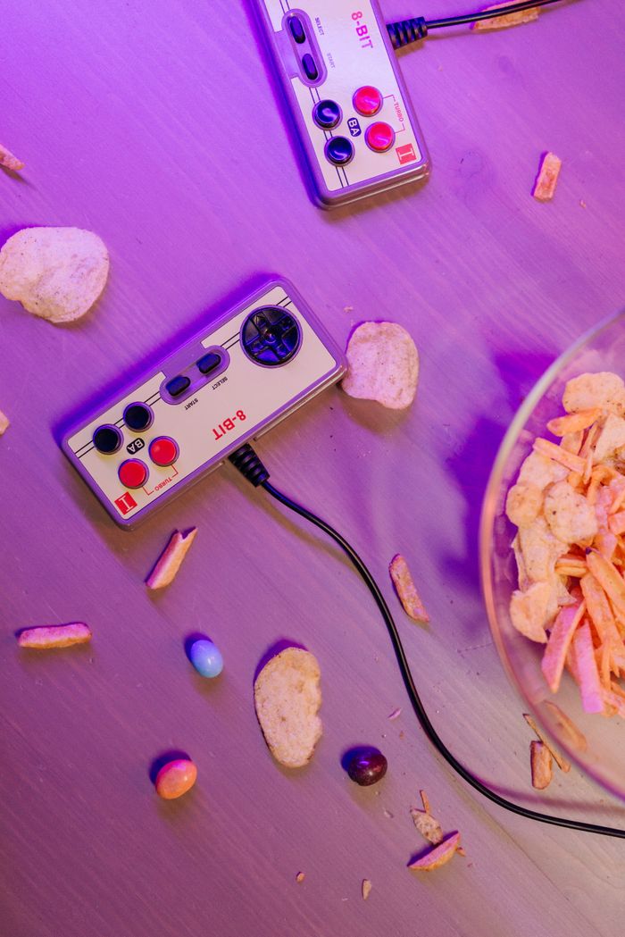 Retro video game controllers with scattered snacks on a table, illuminated by purple lighting.